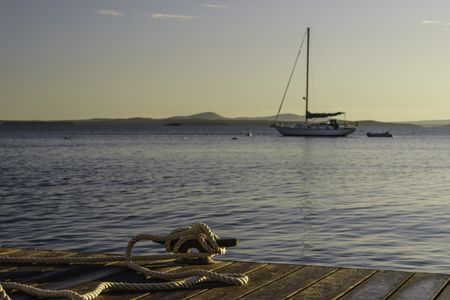 Coastal study in nautical preference: Rope tied to cleat on dock in foreground to secure a boat off camera, while a sailboat lies at anchor beyond, at sunrise in New England (foreground focus)
