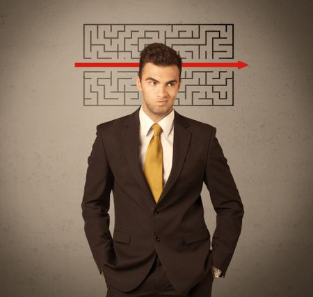 A young handsome business person making facial expression and solving maze with red arrow in front of clear, empty concrete wall background concept