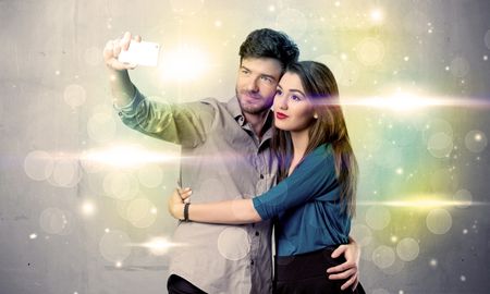 A fresh happy couple taking selfie photo with mobile phone in front of colorful lights glitter wall background concept