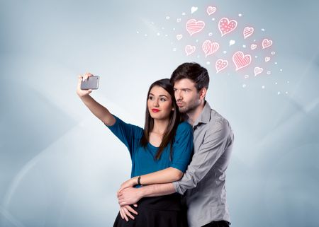 A young couple in love and drawn red hearts taking selfie with a mobile phone in the handsome guy's hand in front of an empty clear grey wall background concept