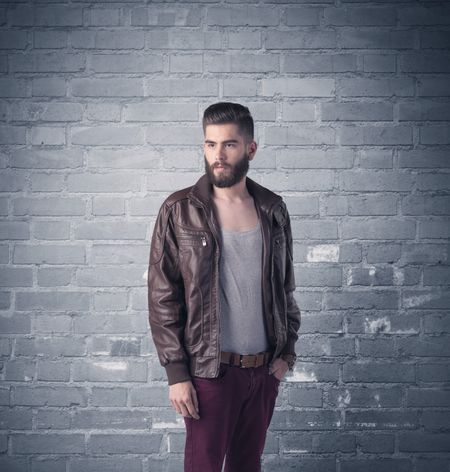 A stylish hipster guy with beard and sunglasses standing in casual clothes in front of an urban blue brick wall background concept