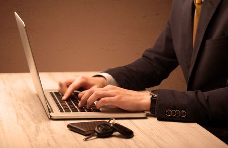 An office worker in elegant suit sitting at desk, typing on portable laptop with empty brown wall background