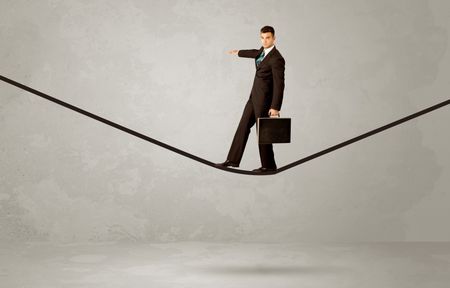 An elegan businessman in suit balancing on a tight rope with a briefcase in front of grey urban wall background environment concept