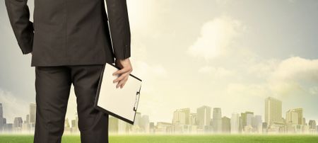 Businessman from the back in front of a city view with clouds and grass