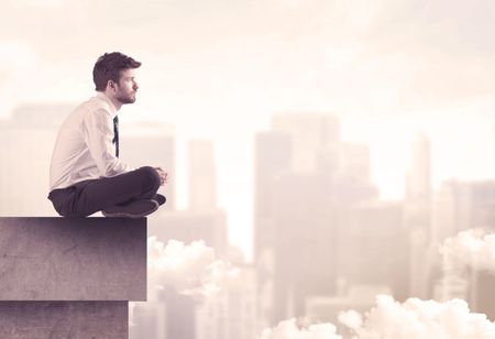 A serious business person sitting with laptop and tablet at the edge of a tall building, looking over cloudy city scape concept