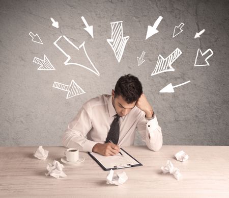 A young businessman sitting at an office desk and working on paperwork with drawn arrows pointing at his head concept 