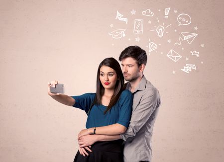 A young couple in love taking selfie with a mobile phone in the handsome guy's hand and drawn media communication icons above them, confused ideas concept