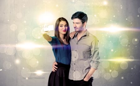 A fresh happy couple taking selfie photo with mobile phone in front of colorful lights glitter wall background concept