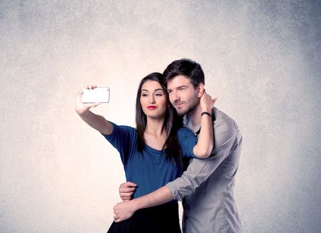 A young couple in love taking selfie with a mobile phone in the handsome guy's hand in front of an empty clear grey wall background concept