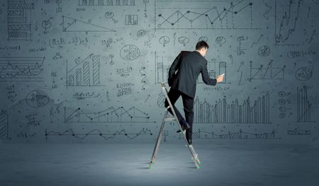 A businessman in modern stylish elegant suit standing on a small ladder and drawing pie and block charts on grey wall background with exponential progressing curves, lines, circles, blocks, numbers