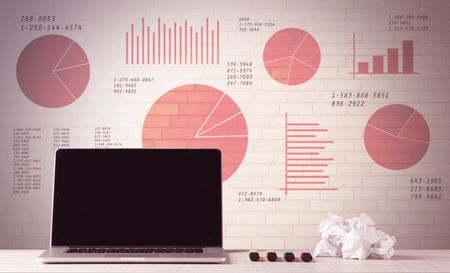 A professional laptop with blank screen sitting on a white office desk in front of wall full of pie charts and graphs concept