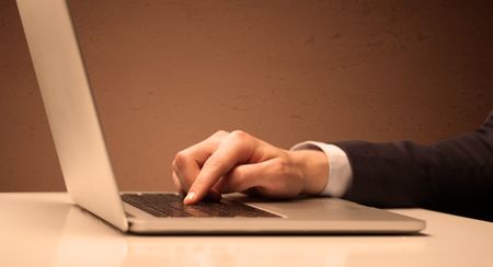 An office worker in elegant suit sitting at desk, typing on portable laptop with empty brown wall background