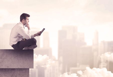 A serious business person sitting with laptop and tablet at the edge of a tall building, looking over cloudy city scape concept
