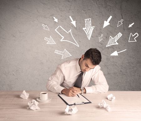 A young businessman sitting at an office desk and working on paperwork with drawn arrows pointing at his head concept 