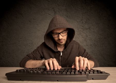 A young hacker with glasses dressed in casual clothes sitting at a desk and working on a computer keyboard in front of black clear concrete wall background concept
