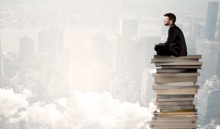 A serious businessman in elegant suit sitting on a pile of giant books in front of a grey city scape with clouds, fog