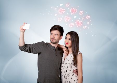 A young couple in love and drawn red hearts taking selfie with a mobile phone in the handsome guy's hand in front of an empty clear grey wall background concept