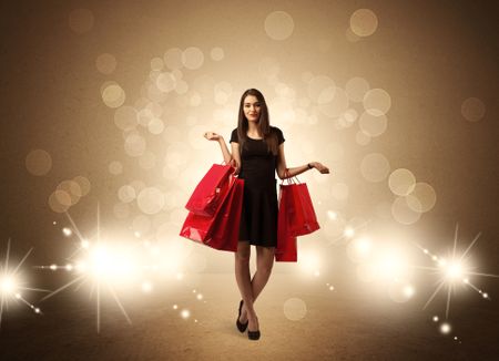 A beautiful elegant woman in black standing with red shopping bags in front of brown background and bright glowing lights concept