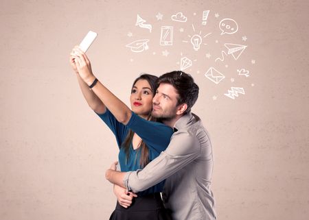 A young couple in love taking selfie with a mobile phone in the handsome guy's hand and drawn media communication icons above them, confused ideas concept