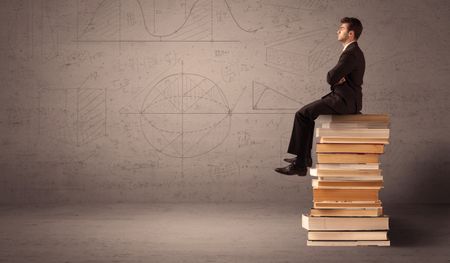 A serious businessman in suit sitting on a pile of giant books in front of a greyish brown wall with drawn lines, angles, numbers, circles and curves.