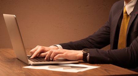An office worker in elegant suit sitting at desk, typing on portable laptop with empty brown wall background