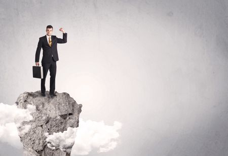 A happy successful businessman standing on a stone cliff with clouds in front of clear empty grey background concept