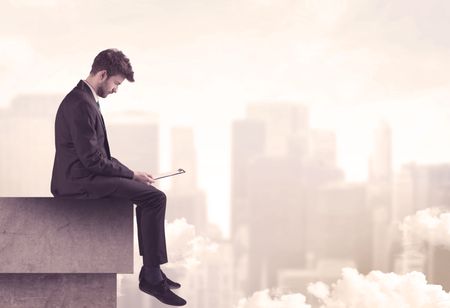 A serious business person sitting with laptop and tablet at the edge of a tall building, looking over cloudy city scape concept