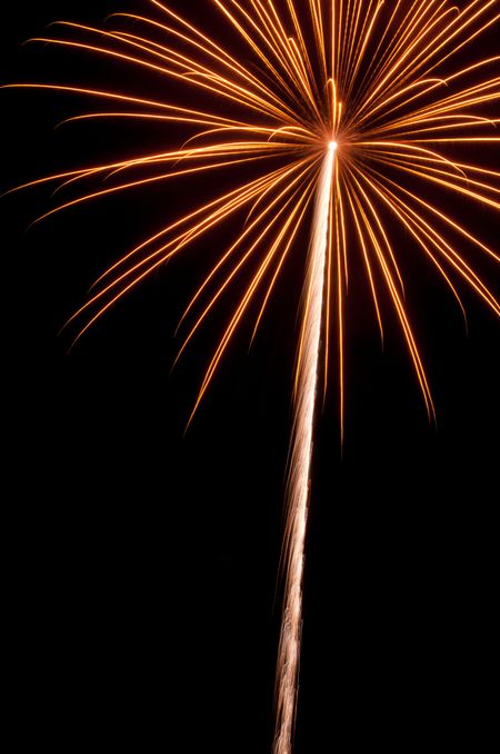 High-altitude burst of yellowish-orange fireworks with long white rocket trail