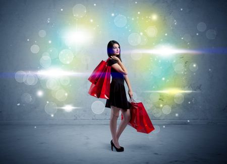 A beautiful young girl standing with long hair and red shopping bags in front of colorful light bokeh urban wall background concept