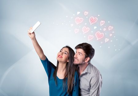 A young couple in love and drawn red hearts taking selfie with a mobile phone in the handsome guy's hand in front of an empty clear grey wall background concept