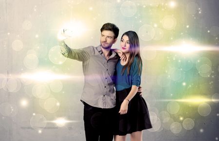 A fresh happy couple taking selfie photo with mobile phone in front of colorful lights glitter wall background concept