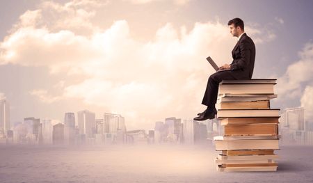 A serious student with laptop tablet in elegant suit sitting on a stack of books in front of cityscape with clouds