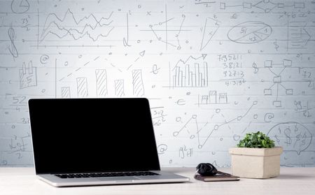 A professional laptop with blank screen sitting on a white office desk in front of wall full of pie charts, calculations and graphs concept