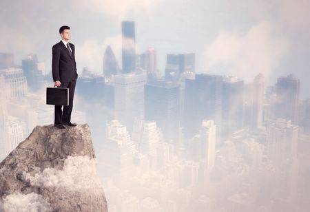 A successful good looking businessman standing on top of a high cliff above the city scape with clouds concept