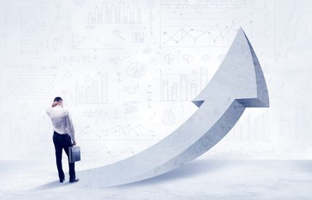 Young sales business person in elegant suit standing with his back in front of a big arrow pointing up and a clear background full of pie charts, numbers