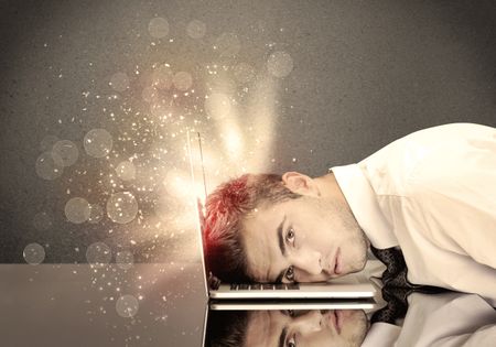 A young depressed business person laying his head on computer keyboard with thoughts exploding from his head illustrated by light beams concept