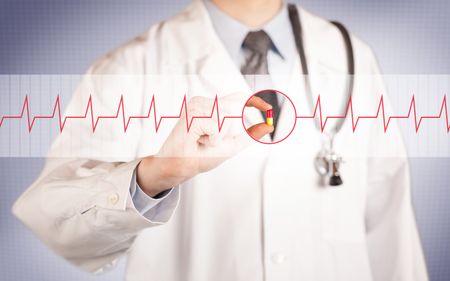 A male doctor in white coat with a stethoscope on one shoulder holding a pill between his fingers focused on a heartbeat graph