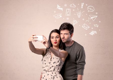 A young couple in love taking selfie with a mobile phone in the handsome guy's hand and drawn media communication icons above them, confused ideas concept