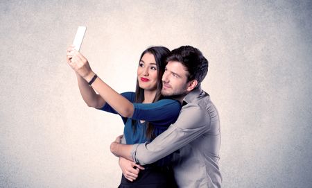 A young couple in love taking selfie with a mobile phone in the handsome guy's hand in front of an empty clear grey wall background concept