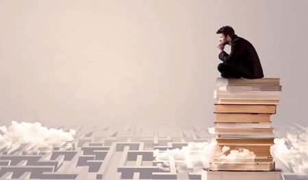 A serious businessman in suit sitting on a pile of giant books in front of a grey wall with clouds, labirynth