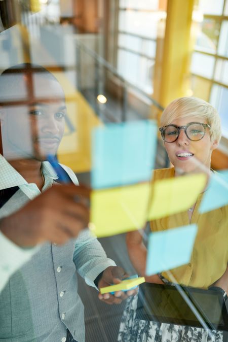 Two creative millenial small business owners working on social media strategy brainstorming using adhesive notes in windows