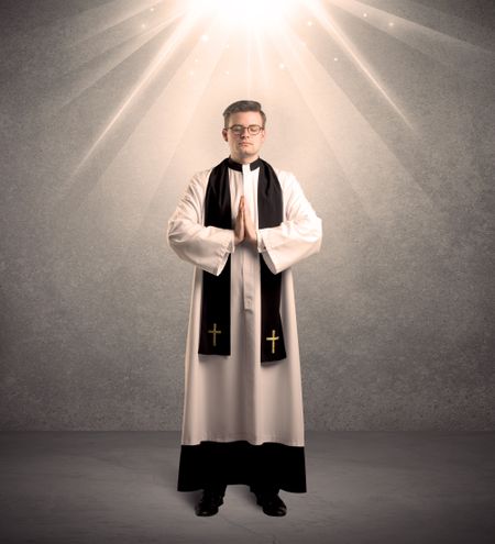 A male religious young priest in black and white dress giving his blessing, holding the holy bible while being illuminated from strong light beams coming from above concept