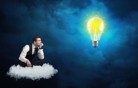 Caucasian businessman sitting on a white fluffy cloud lookind and wondering at a big, shiny, glowing yellow lightbulb