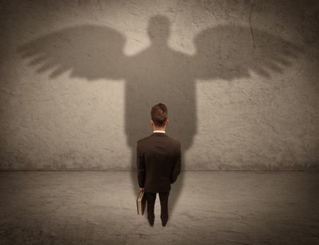 A successful businessman holding a briefcase is giving thumbs up for his angel shadow in front of a wall concept