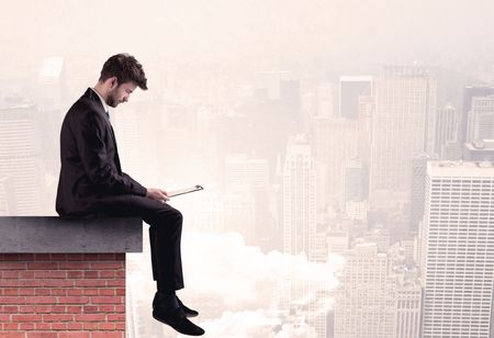 An elegant businessman in modern suit sitting on the top of a brick building, looking over the cityscape with clouds concept