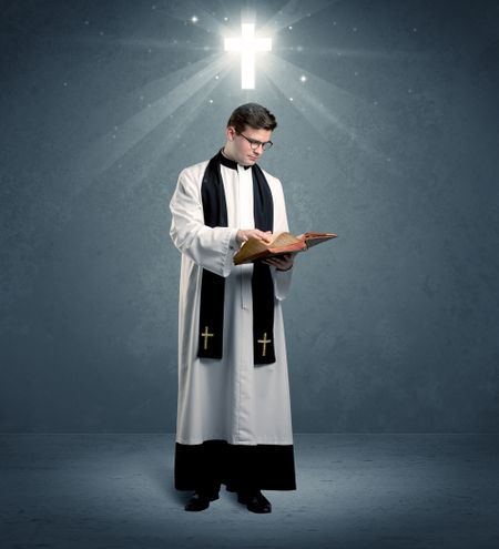 A young caucasian priest with deep faith blessing with the holy bible in his hand in front of blue wall background with illustrated glowing cross concept.