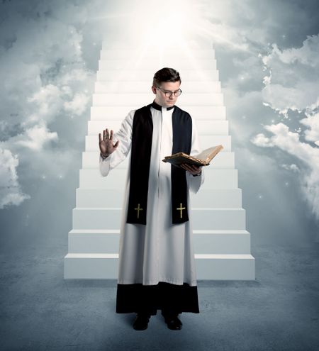 A young religious happy priest standing in front of the stairway to heaven concept with clouds and bright lights coming from above.