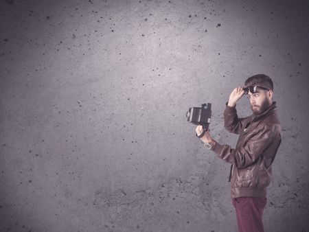 A stylish funny hipster person holding a vintage camera and taking photographs in front of a concrete clear empty urban wall background concept