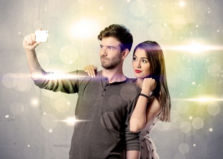 A fresh happy couple taking selfie photo with mobile phone in front of colorful lights glitter wall background concept