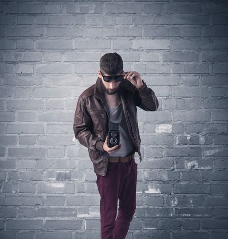 A stylish hipster guy with beard and sunglasses standing in casual clothes in front of an urban blue brick wall background concept